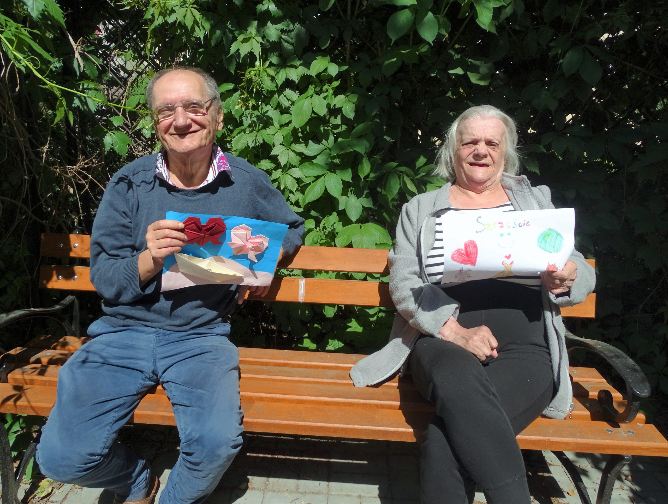 uśmiechnięci seniorzy siedzą na ławce i pokazują karty z pozdrowieniami od młodzieży