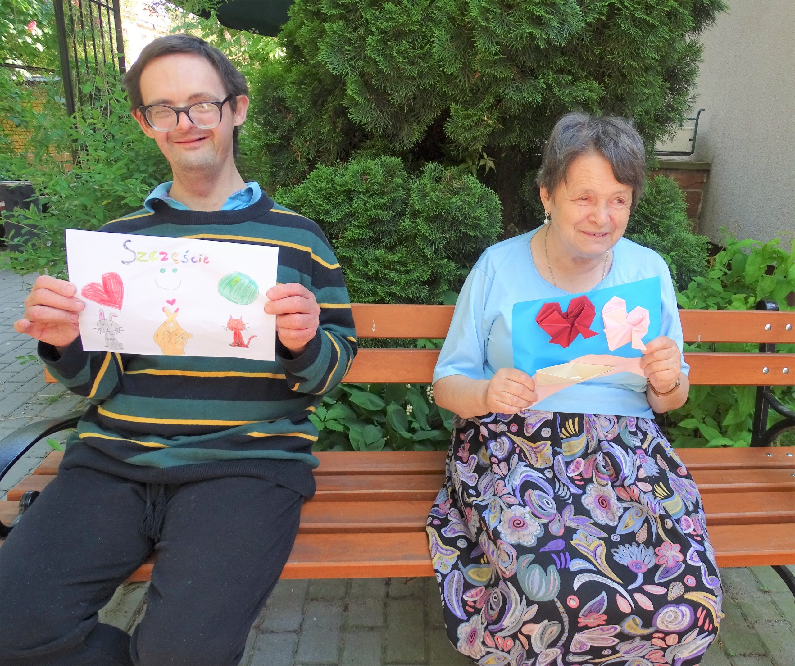 Dwoje uśmiechniętych Mieszkańców Domu siedzi na ławce i pokazuje karty z pozdrowieniami od młodzieży