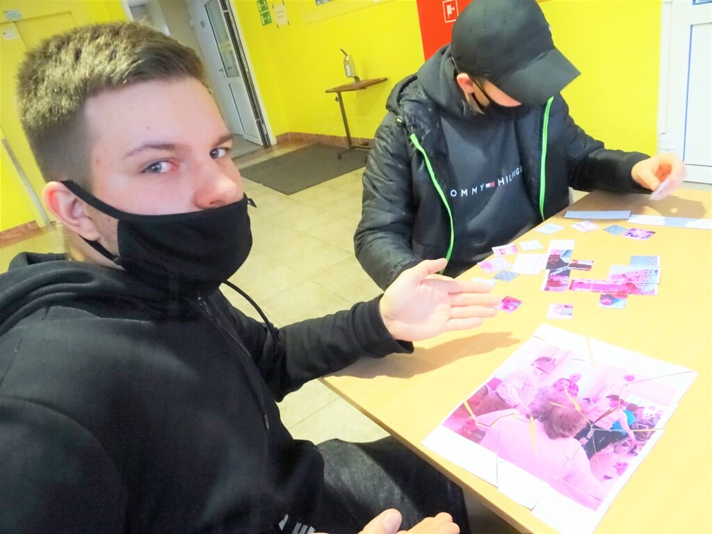 Na pierwszym planie młody mężczyzna pokazujący ułożone puzzle, w tle drugi mężczyzna układający puzzle