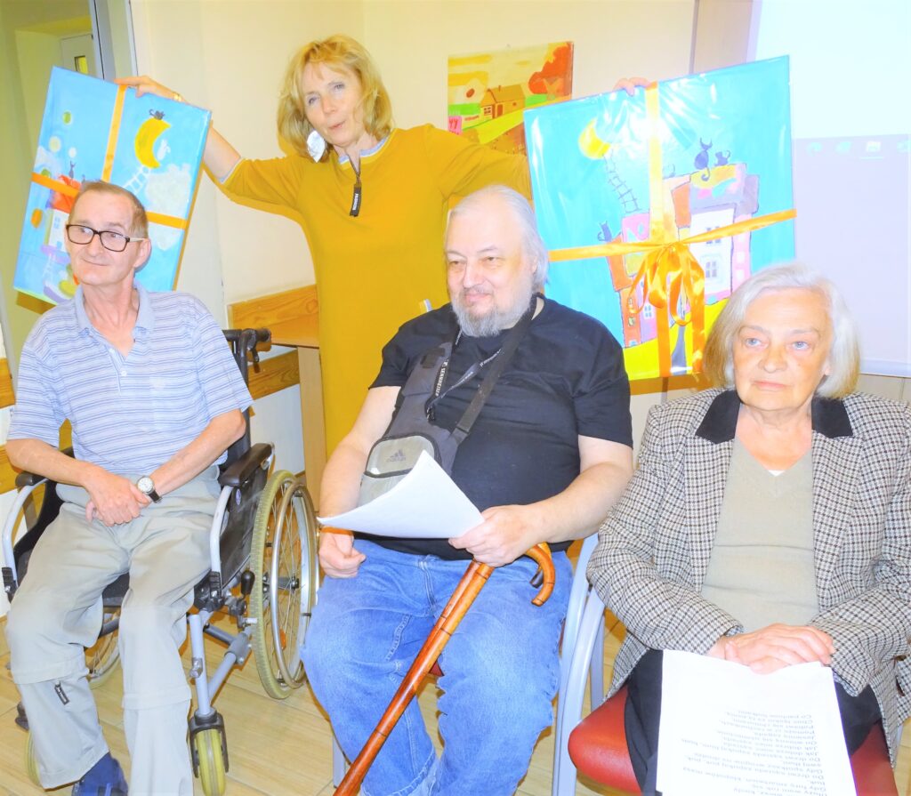 Seniorzy wraz z kobietą trzymającą dwa własnoręcznie namalowane obrazy